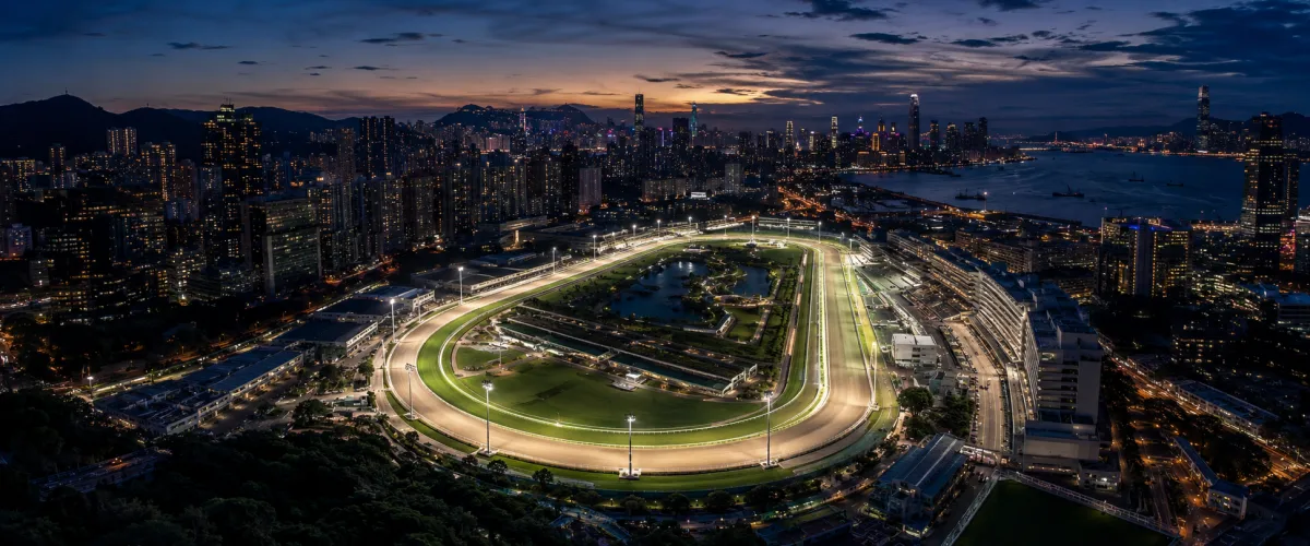 Aerial view of a floodlit racecourse within a coastal city skyline at dusk